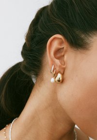 Goldene Creolen mit glänzender Oberfläche, die eine hängende weiße Perle und einen kleinen klaren Kristallakzent aufweisen. Glatte, abgerundete Formen.