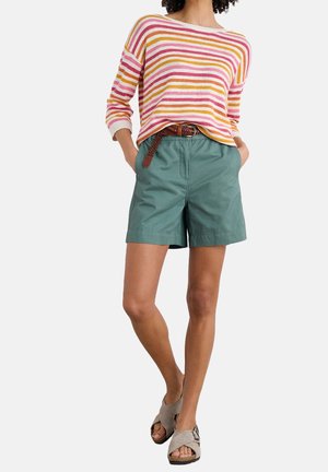 Femme portant un pull rayé rose, orange et blanc, un short vert avec une ceinture marron et des sandales beiges, debout les mains dans les poches.