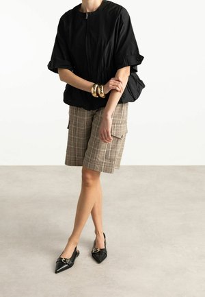 Persoon draagt een zwarte top met rits, geruite cargoshorts tot op de knie, zwarte puntige slingbackhakken en gouden armbanden aan één arm.