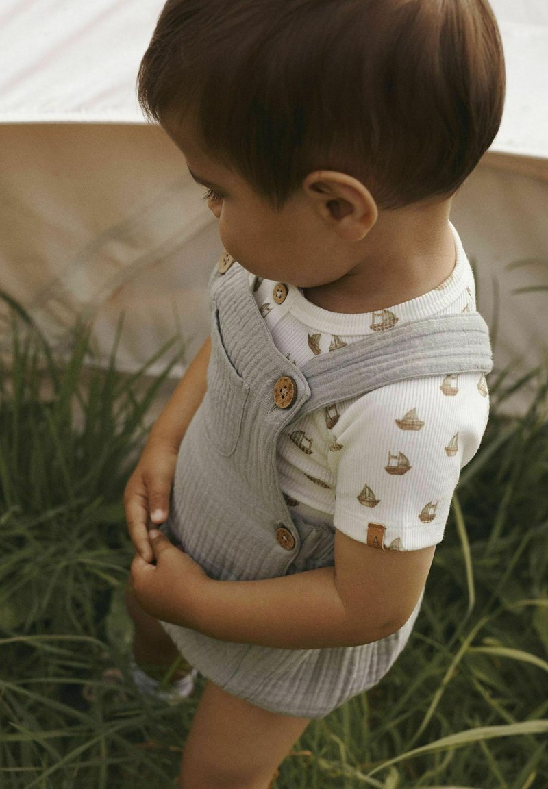 Grå tygoverall med träknappar, bärs över en vit ribbad tröja med små båt-mönster. Bakgrunden innehåller grön gräs.