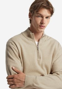 Beige halvmaskad tröja i mjukt material, med långa ärmar, en ribbad krage och en slät yta.