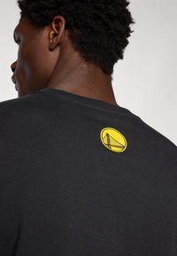 Svart T-skjorte med et rundt gult logo som har en bro-design på øvre del av ryggen. Bomullsstoff med en jevn tekstur. Klassisk passform.