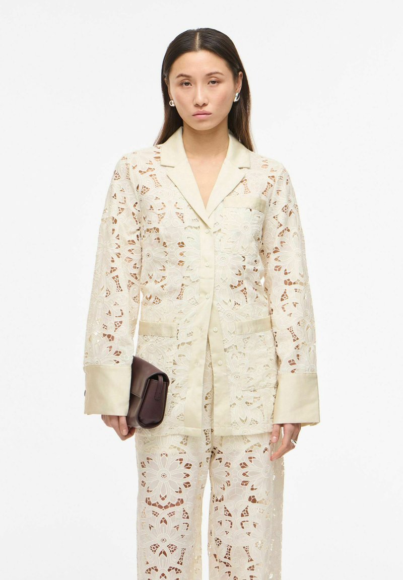 Femme portant un costume en dentelle crème transparente avec des motifs floraux, tenant une pochette marron foncé, debout devant un fond blanc uni.