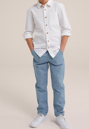 Chłopiec w białej koszuli na guziki z podwiniętymi rękawami, niebieskich spodniach i białych tenisówkach, stojący z rękami w kieszeniach.