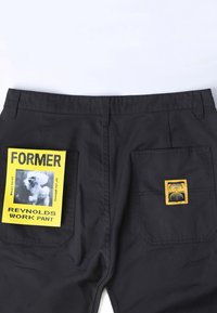 Pantalon de travail noir avec de grandes poches arrière ornées d'un patch jaune représentant une image de chien et un texte, ainsi qu'un petit logo jaune sur la poche droite.