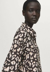 Srajca s ovratnikom, ki ima rjavo, črno in kremasto abstraktno pikčasto vzorec, izdelana je iz lahkega blaga z gladko teksturo.