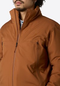 Mann trägt eine braune Jacke mit hohem Kragen und Reißverschluss, die zwei Reißverschlusstaschen auf der Brust sowie ein dezentes Markenlogo auf der linken Seite hat.