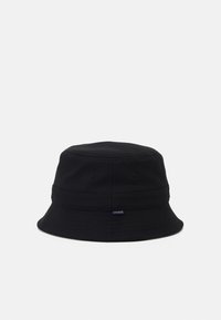 Svart bøttehatt laget av teksturert stoff. Har en bred, fleksibel skygge og et lite Lacoste-logo merke på siden.