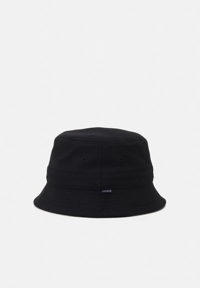 Chapeau seau noir en tissu texturé. Il dispose d'un large bord flexible et d'une petite étiquette logo Lacoste sur le côté.