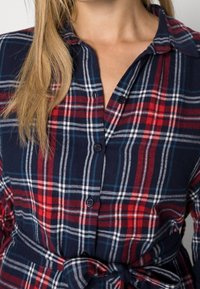 Camicia a quadri con una combinazione di colori blu navy, rosso e bianco. Realizzata in un tessuto morbido con un'apertura frontale con bottoni e dettagli sul punto vita annodati.