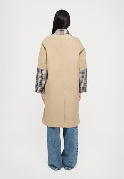 Beige coat with a checkered pattern on the collar and sleeves, featuring a relaxed design and long, wide sleeves. Worn over blue jeans.