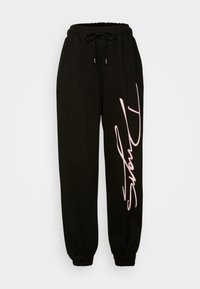 Svarta joggingsbyxor i mjukt tyg med elastiskt midjeband och dragsko. Har en rosa kursiv logotyp på vänster ben. Avsmalnande vid anklarna.
