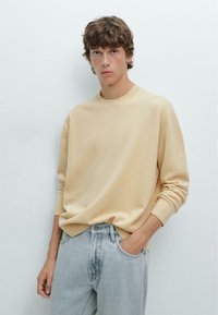 Light beige sweater in soft knit fabric, round neck, and ribbed cuffs. Paired with light blue denim jeans. Subtle design, casual style.