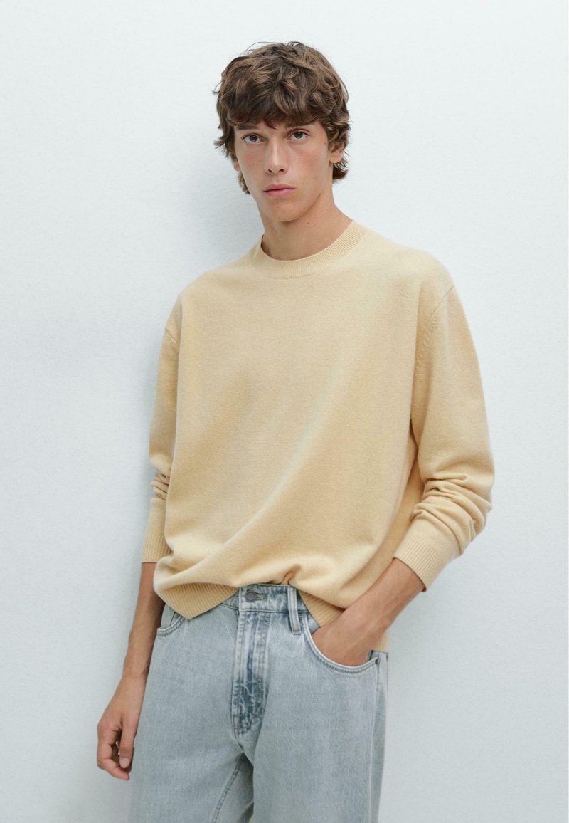 Light beige sweater in soft knit fabric, round neck, and ribbed cuffs. Paired with light blue denim jeans. Subtle design, casual style.