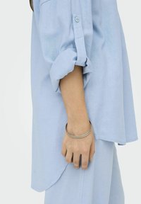 Chemise bleu clair à manches retroussées en tissu doux, dotée d'un poignet à boutons. Bracelet en argent au poignet, avec des détails de texture subtils.