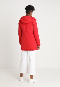 Rote Parka mit Kapuze, glatter Textur, langen Ärmeln und knielanger Länge. Verfügt über eine Kordelzugkapuze und goldfarbene Hardware-Akzente.