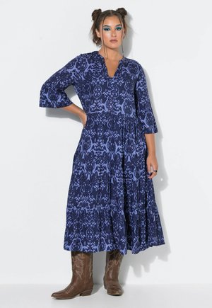 Donna che indossa un vestito midi blu con fantasia, maniche a tre quarti e stivali da cowboy marroni, in piedi davanti a uno sfondo bianco.
