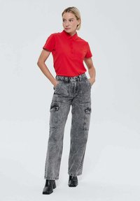 Femme aux cheveux courts blonds, portant un polo rouge, un jean cargo gris taille haute et des bottines noires, se tenant les mains dans les poches.