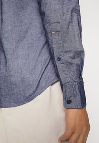 Bras droit portant une chemise bleue à manches longues avec un poignet boutonné, tenu par une main près d’un pantalon beige.