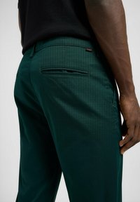 Pantaloni verdi con una texture liscia e un sottile motivo a quadri. Presenta tasche posteriori e un'etichetta del marchio cucita sulla vita.