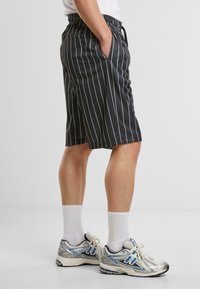 Shorts noirs avec des rayures verticales blanches, taille élastique, poches latérales, longueur genou. Associés à des chaussures de sport argentées et des chaussettes blanches.