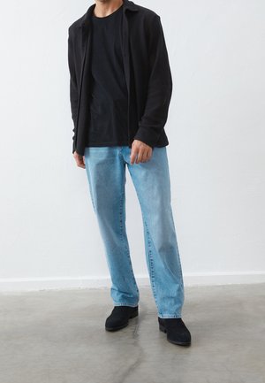 Veste noire texturée portée sur un t-shirt noir, associée à un jean bleu clair et des bottes en daim noir, debout sur un sol en béton.