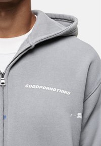 Felpa grigia con zip realizzata in tessuto morbido, con logo bianco sul petto e cappuccio strutturato. Gli accenti includono una piccola macchia blu.