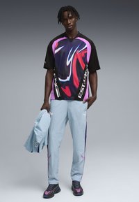 Νεαρός άνδρας που φοράει πουκάμισο με κοντά μανίκια και αφηρημένο τύπωμα σε μαύρο, μοβ και κόκκινο, ανοιχτόχρωμο μπλε παντελόνι φόρμας και κρατάει ένα ταιριαστό ανοιχτόχρωμο μπλε σακάκι.