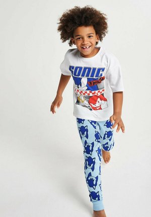 Smiling child with curly hair wearing Sonic the Hedgehog graphic t-shirt and blue pants with Sonic faces, walking barefoot on plain background.