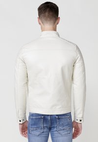 Chaqueta de cuero blanca con un diseño ajustado, acentos acolchados en los hombros y puños con botones a presión, combinada con jeans azules, vista desde la parte de atrás.
