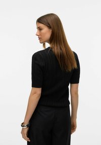 Černý svetr s krátkým rukávem s texturovaným vzorem a žebrovanými detaily, spárovaný s černými kalhotami a metalickým náramkem.