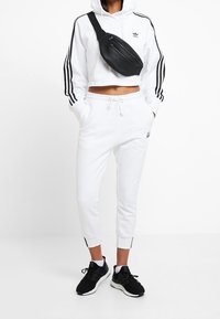 Bílá zkrácená mikina s černými pruhy, bílé tepláky a černé sportovní boty. Navíc je přes tělo připevněn černý ledvinkový batoh.