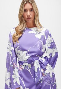 Lila Kleid mit langen Ärmeln, das ein florales Muster in Weiß und Pink aufweist. Mit einem gebundenen Taillen-Detail und glatter Stoffstruktur.