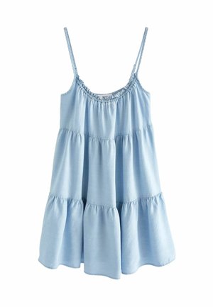 Femme portant une robe bleu clair sans manches à plusieurs volants, debout près d'un mur avec la lumière du soleil et des ombres en arrière-plan.