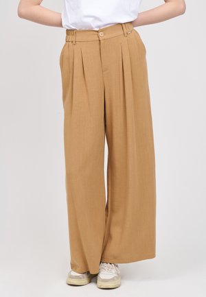 Frau trägt hochtaillierte, weit geschnittene Kamelhosen kombiniert mit weißen Turnschuhen und einem weißen, hineingesteckten Hemd.