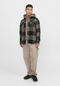 Camisa de franela a cuadros en negro y verde sobre una sudadera beige, combinada con pantalones beige y calzado negro. Presenta bolsillos con botones y cordón ajustable.