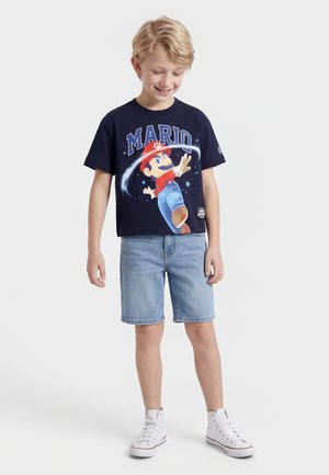 Ξανθός αγόρι που φοράει μπλε σκούρο μπλουζάκι με γραφικό του Μάριο, ανοιχτόχρωμα τζιν σορτς και λευκά πάνινα παπούτσια, χαμογελώντας και κοιτάζοντας προς τα κάτω.