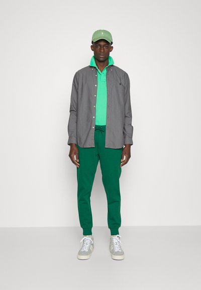 Camisa de botones gris sobre un polo verde, combinada con pantalones de chándal verdes y zapatillas gris, rematada con una gorra verde claro.