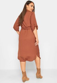 Braunes, mittellanges Kleid mit einem gegurteten Bund, aufgerollten Ärmeln und einem geschwungenen Saum. Kombiniert mit hellbraunen, gerippten Ankle Boots. Glatte Textur.