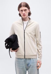 Νεαρός άνδρας με μεσαίου μήκους μαλλιά φοράει μπεζ ζακέτα με φερμουάρ και ανοιχτόχρωμο τζιν παντελόνι, κρατώντας ένα μαύρο διπλωμένο σακίδιο στον αριστερό του χέρι.