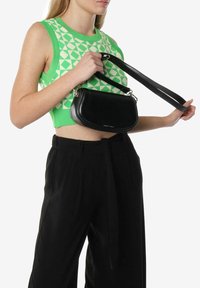 Borsa in pelle nera con forma curvata, tracolla staccabile e hardware in metallo. La borsa è tenuta da una persona che indossa un top verde con fantasia.