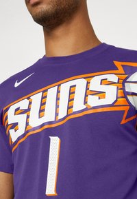 Lilla basketballtrøje med "SUNS" i hvid og orange, med perforeret nummer "1." Fremstillet af åndbart stof. Nike-logo til stede.