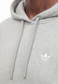 Gros plan d'une personne portant un sweat à capuche gris clair avec cordons de serrage et un petit logo Adidas trefoil blanc sur la poitrine.