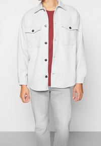 Lys grå knap-up skjorte med to brystlommer, rund kant og mørke knapper, lagdelt over en bordeaux crew-neck skjorte og lys grå jeans.