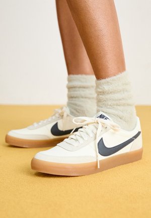 Nogi w białych tenisówkach Nike z czarnym logo i gumowymi podeszwami, w towarzystwie puszystych białych skarpet, stojące na żółtej powierzchni.