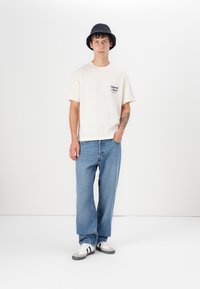 T-shirt crème à manches courtes avec logo sur la poitrine, associé à un jeans droit bleu clair et des baskets blanches avec des accents noirs. Chapeau seau noir.