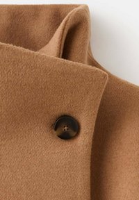 Detalle de un abrigo de lana marrón que presenta una tela suave y texturizada, cuello doblado y un botón marrón oscuro con cuatro agujeros.