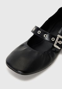 Zapato de ballet de cuero negro con punta cuadrada, que presenta una correa con herrajes plateados y ojetes para un detalle adicional. Textura suave, diseño de calce fácil.