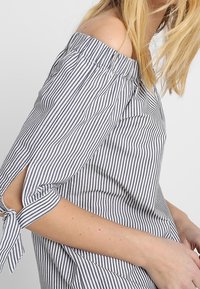 Off-shoulder randig skjorta i blått och vitt, med korta ärmar med knytdetaljer och en avslappnad passform. Tillverkad av lättviktig tyg.