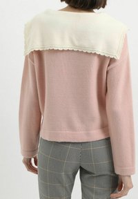 Persona vestida con un suéter de punto rosa con cuello de chal crema y pantalones a cuadros en blanco y negro, vista desde atrás contra un fondo neutro.